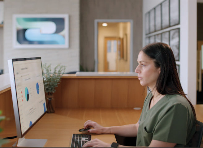 A receptionist sits at a front desk of a dental office and uses Link AI for A-dec+ software on a computer